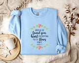 A light blue sweatshirt with a printed Bible verse and a floral design, along with a mug and a branch of cotton flowers, all placed on a white fur rug.