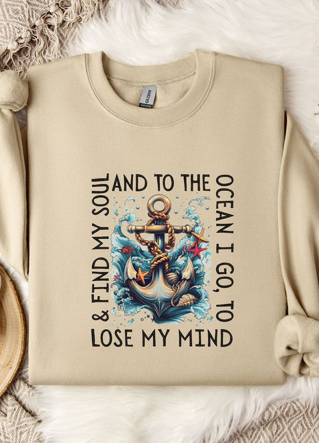 a beige sweatshirt with a graphic design of an anchor and waves, along with the text "& FIND MY SOUL AND TO THE OCEAN I GO TO LOSE MY MIND". The sweatshirt is laid out on a bed with a white blanket, and there are some decorative elements such as a snowflake and a branch with cotton balls.