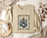 a beige sweatshirt with a graphic design of an anchor and waves, along with the text "& FIND MY SOUL AND TO THE OCEAN I GO TO LOSE MY MIND". The sweatshirt is laid out on a bed with a white blanket, and there are some decorative elements such as a snowflake and a branch with cotton balls.