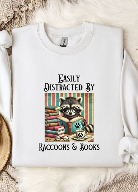 a white t-shirt with a graphic design of a raccoon reading books, accompanied by the text "Easily Distracted By Raccoons & Books".