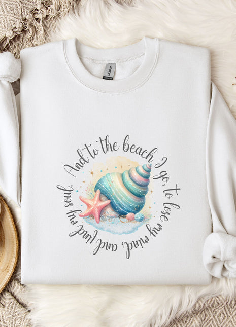 A white sweatshirt with a colorful graphic of a seashell and starfish, along with the text "And to the beach go, to lose and to love again" printed on it, is displayed on a bed with a white blanket and a mug.