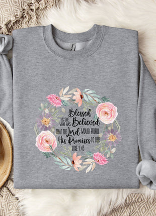 A gray sweatshirt with a floral design and text printed on it, along with a mug, a branch of cotton bolls, and a blanket on a bed.