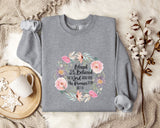 A gray sweatshirt with a floral design and text printed on it, along with a mug, a branch of cotton bolls, and a blanket on a bed.