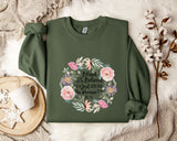 A green sweatshirt with a floral design and text printed on it, along with a mug, a white mug, and some white flowers on a white surface.