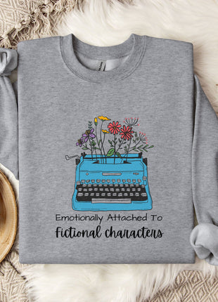 A gray sweatshirt with a graphic design of a typewriter filled with colorful flowers, accompanied by the text "Emotionally Attached To Fictional Characters".