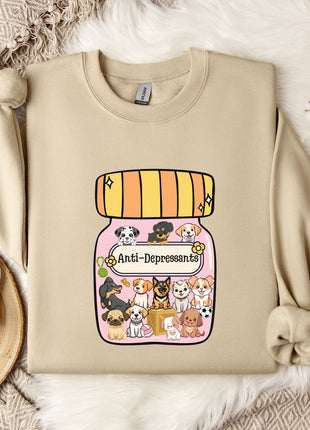 A beige sweatshirt with a graphic design of a jar containing various cartoon animals, including dogs, cats, and birds, along with the text "Ani-Depressants". The sweatshirt is placed on a white fur rug, and there is a mug and some cotton balls nearby.