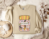 A beige sweatshirt with a graphic design of a jar containing various cartoon animals, including dogs, cats, and birds, along with the text "Ani-Depressants". The sweatshirt is placed on a white fur rug, and there is a mug and some cotton balls nearby.