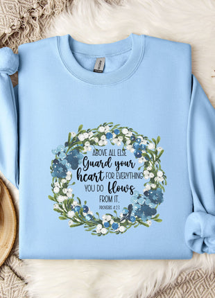A light blue sweatshirt with a floral wreath design and a quote printed on it, along with a mug and some white flowers on a bed.