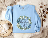 A light blue sweatshirt with a floral wreath design and a quote printed on it, along with a mug and some white flowers on a bed.