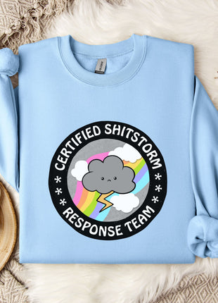 a light blue sweatshirt with a graphic design on it, placed on a bed with a white blanket and a mug.