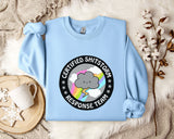 a light blue sweatshirt with a graphic design on it, placed on a bed with a white blanket and a mug.