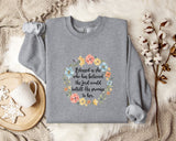 A gray sweatshirt with a floral design and text printed on it, placed on a bed with a white blanket and a white mug.