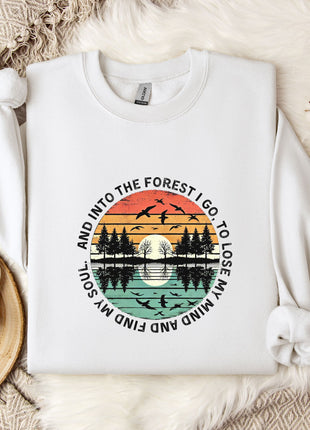 A white sweatshirt with a graphic design depicting a sunset over a forest, surrounded by silhouetted trees and birds. The text around the design reads "And into the forest I go, to lose my soul and find my mind".
