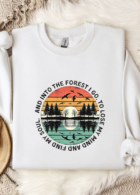 A white sweatshirt with a graphic design depicting a sunset over a forest, surrounded by silhouetted trees and birds. The text around the design reads "And into the forest I go, to lose my soul and find my mind".