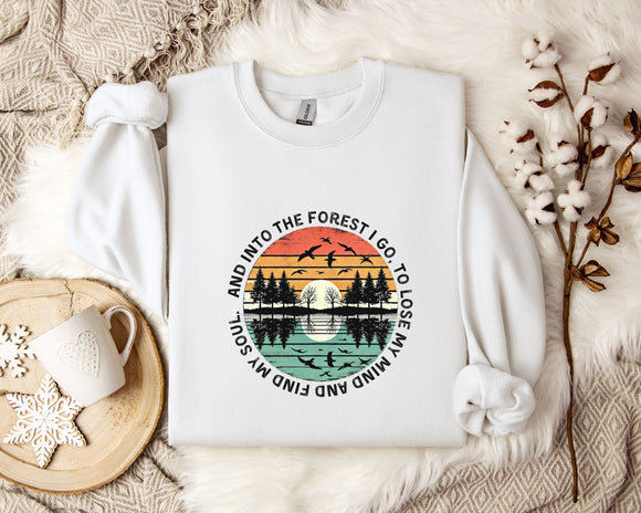 A white sweatshirt with a graphic design depicting a sunset over a forest, surrounded by silhouetted trees and birds. The text around the design reads 