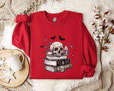 a red sweater with a skull and bat design, along with a stack of books and a mug, all placed on a white fur rug.