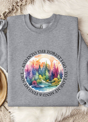 A gray sweatshirt with a colorful forest scene printed on it, along with a mug, a branch of cotton flowers, and a blanket on a fur-lined surface.