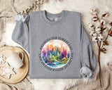 A gray sweatshirt with a colorful forest scene printed on it, along with a mug, a branch of cotton flowers, and a blanket on a fur-lined surface.