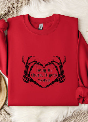 A red sweater with a heart-shaped graphic and the text "hang in there, it gets worse" printed on it, along with a mug and some white flowers on a white surface.