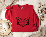 A red sweater with a heart-shaped graphic and the text "hang in there, it gets worse" printed on it, along with a mug and some white flowers on a white surface.
