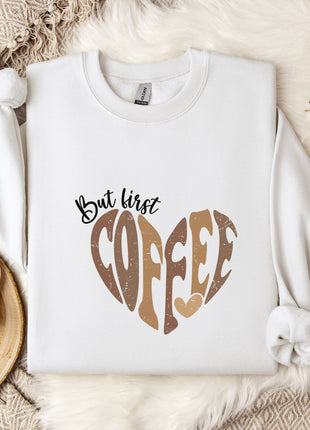 A white sweatshirt with a heart-shaped design and the text "But first coffee" printed on it, along with a mug and some flowers on a bed.