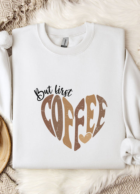 A white sweatshirt with a heart-shaped design and the text "But first coffee" printed on it, along with a mug and some flowers on a bed.