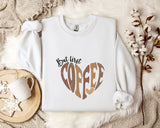 A white sweatshirt with a heart-shaped design and the text "But first coffee" printed on it, along with a mug and some flowers on a bed.