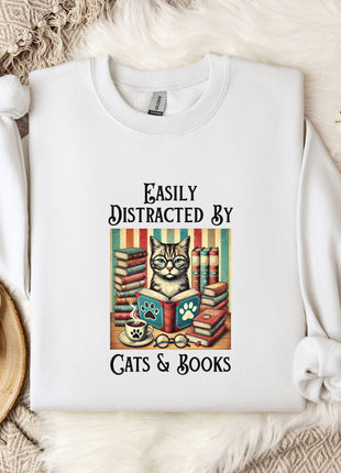 A white t-shirt with a graphic design of a cat surrounded by books and a cup of coffee, along with the text "Easily Distracted By Cats & Books".