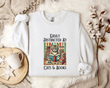 A white t-shirt with a graphic design of a cat surrounded by books and a cup of coffee, along with the text "Easily Distracted By Cats & Books".