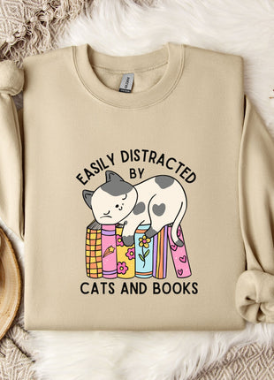 a beige sweatshirt with a graphic design of a cat sitting on a stack of books, accompanied by the text "EASILY DISTracted BY CATS AND BOOKS". The sweatshirt is laid out on a bed with a white blanket, and there are some books and a mug nearby.