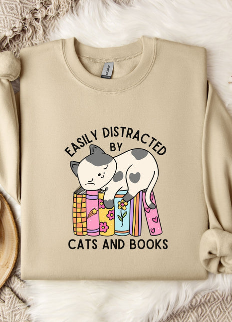 a beige sweatshirt with a graphic design of a cat sitting on a stack of books, accompanied by the text "EASILY DISTracted BY CATS AND BOOKS". The sweatshirt is laid out on a bed with a white blanket, and there are some books and a mug nearby.