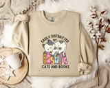 a beige sweatshirt with a graphic design of a cat sitting on a stack of books, accompanied by the text "EASILY DISTracted BY CATS AND BOOKS". The sweatshirt is laid out on a bed with a white blanket, and there are some books and a mug nearby.