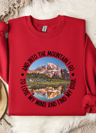 A red sweater with a mountain landscape design and the text "AND INTO THE MOUNTAIN I GO TO LOSE MY MIND AND FIND MY SOUL" printed on it, along with a mug and some white flowers.