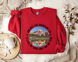 A red sweater with a mountain landscape design and the text "AND INTO THE MOUNTAIN I GO TO LOSE MY MIND AND FIND MY SOUL" printed on it, along with a mug and some white flowers.