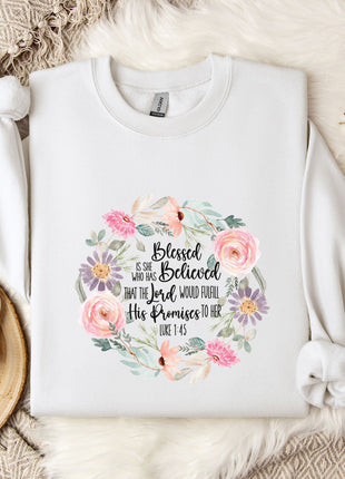 A white sweatshirt with a floral design and text printed on it, placed on a bed with a white blanket and a white mug.