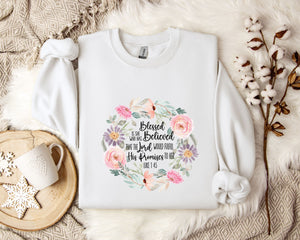 A white sweatshirt with a floral design and text printed on it, placed on a bed with a white blanket and a white mug.