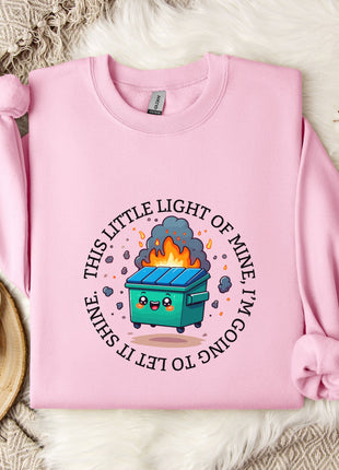 a pink sweatshirt with a graphic design depicting a fire-breathing trash can, surrounded by a circular text that reads "This little light of mine shines bright".