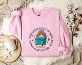 a pink sweatshirt with a graphic design depicting a fire-breathing trash can, surrounded by a circular text that reads "This little light of mine shines bright".