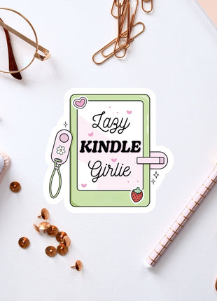 a sticker with the text "Lazy Kindle Girlie" surrounded by various office supplies such as a pen, a pair of glasses, and a notebook.