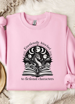 A pink sweatshirt with a graphic design of a dragon and moon, along with the text "Emotionally Attached to Fiction" printed on it, is displayed on a bed with a white blanket and a mug.