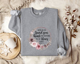 a gray sweatshirt with a floral design and text printed on it, placed on a bed with a white blanket and a mug.