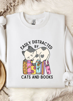 a white t-shirt with a graphic design of a cat sitting on a stack of books, accompanied by the text "EASILY DISTracted BY CATS AND BOOKS". The t-shirt is laid out on a bed with a blanket and a mug, and there are some flowers and a branch nearby.