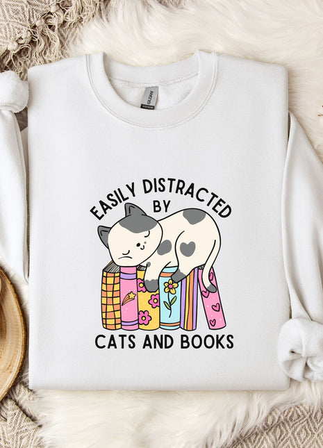 a white t-shirt with a graphic design of a cat sitting on a stack of books, accompanied by the text "EASILY DISTracted BY CATS AND BOOKS". The t-shirt is laid out on a bed with a blanket and a mug, and there are some flowers and a branch nearby.