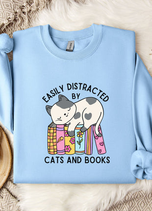 A light blue sweatshirt with a cartoon cat and the text "EASILY DISTracted BY CATS AND BOOKS" printed on it, along with a mug and some flowers.