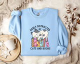 A light blue sweatshirt with a cartoon cat and the text "EASILY DISTracted BY CATS AND BOOKS" printed on it, along with a mug and some flowers.