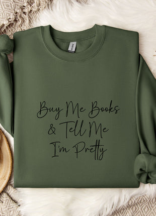 A green sweatshirt with the text "Buy Me Books & Tell Me I'm Pretty" printed on it, along with a mug and some flowers on a bed.