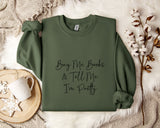A green sweatshirt with the text "Buy Me Books & Tell Me I'm Pretty" printed on it, along with a mug and some flowers on a bed.