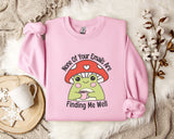 a pink sweatshirt with a mushroom character and text, placed on a bed with a white blanket and a mug.