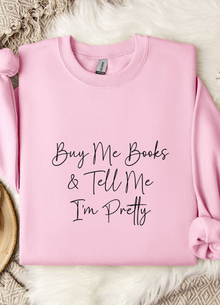 A pink sweatshirt with the text "Buy Me Books & Tell Me I'm Pretty" printed on it, along with a mug and some flowers on a bed.