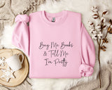 A pink sweatshirt with the text "Buy Me Books & Tell Me I'm Pretty" printed on it, along with a mug and some flowers on a bed.